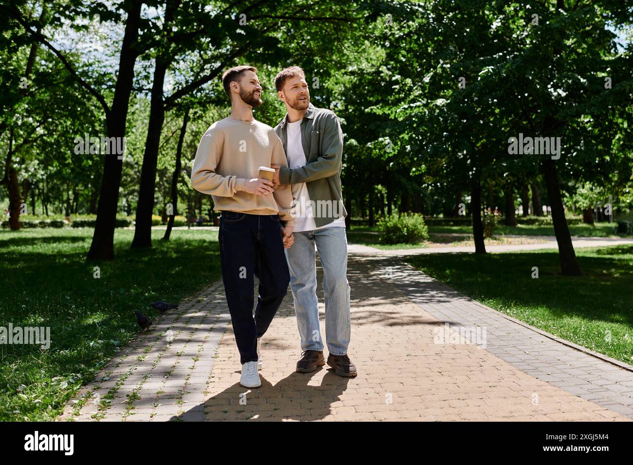 Due uomini barbuti, vestiti in modo informale, si tengono per mano e attraversano un parco in una giornata di sole. Foto Stock