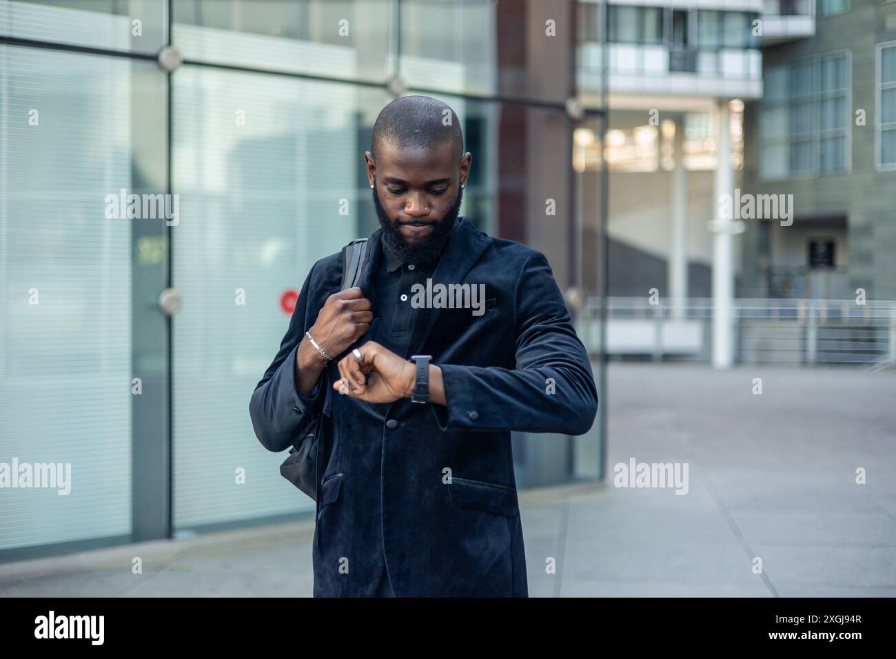 Uomo d'affari nero africano in costume e con uno zaino che controlla l'ora sul suo orologio da polso con un'espressione scontenta e frustrata. La truffa della scena Foto Stock