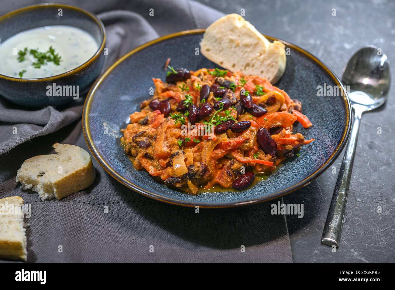 Fagioli rossi, stufato con peperoni, pomodori e cipolle serviti con una salsa di yogurt in ciotole blu su sfondo ardesia, di stile vegetariano levante Foto Stock