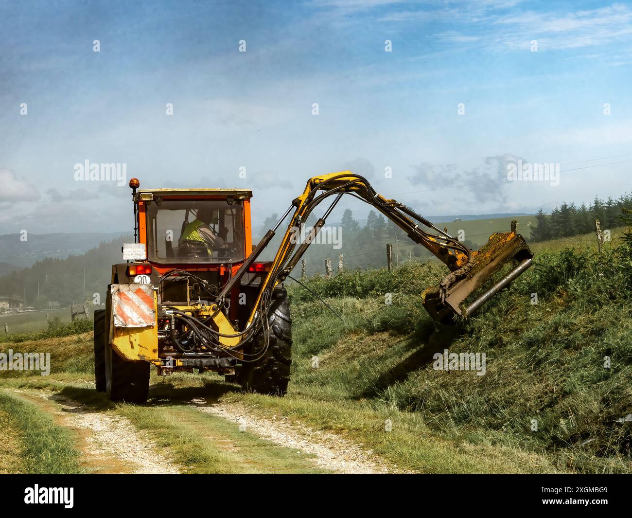 Un trattore con un'attrezzatura a lama rotante taglia una collina, lasciando un'andana di erba tagliata sulla sua scia. Il trattore procede su un percorso sterrato con una vista o. Foto Stock