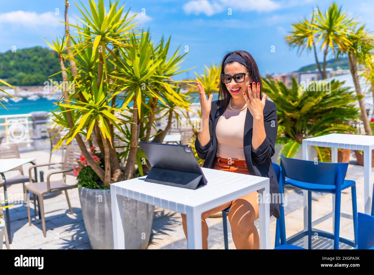 Elegante donna d'affari di bellezza che celebra le notizie durante le riunioni online utilizzando un tablet digitale da una lussuosa terrazza costiera con vista sul mare Foto Stock