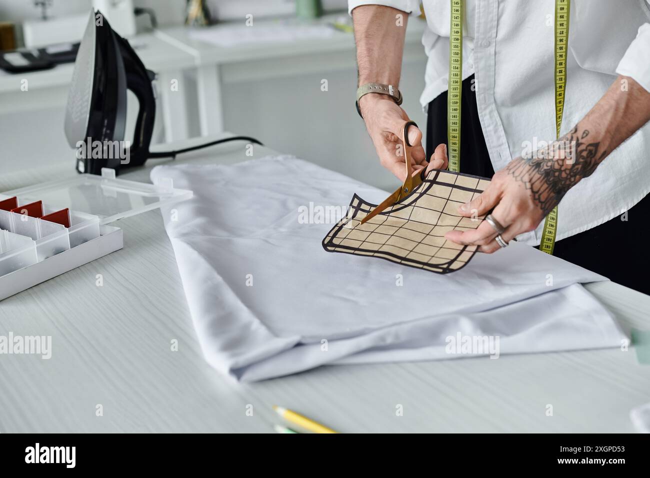 Un giovane uomo, con una camicia bianca e un metro a nastro, taglia il tessuto nel suo atelier. È focalizzato sulla sostenibilità e sul rilancio dell'abbigliamento vecchio. Foto Stock