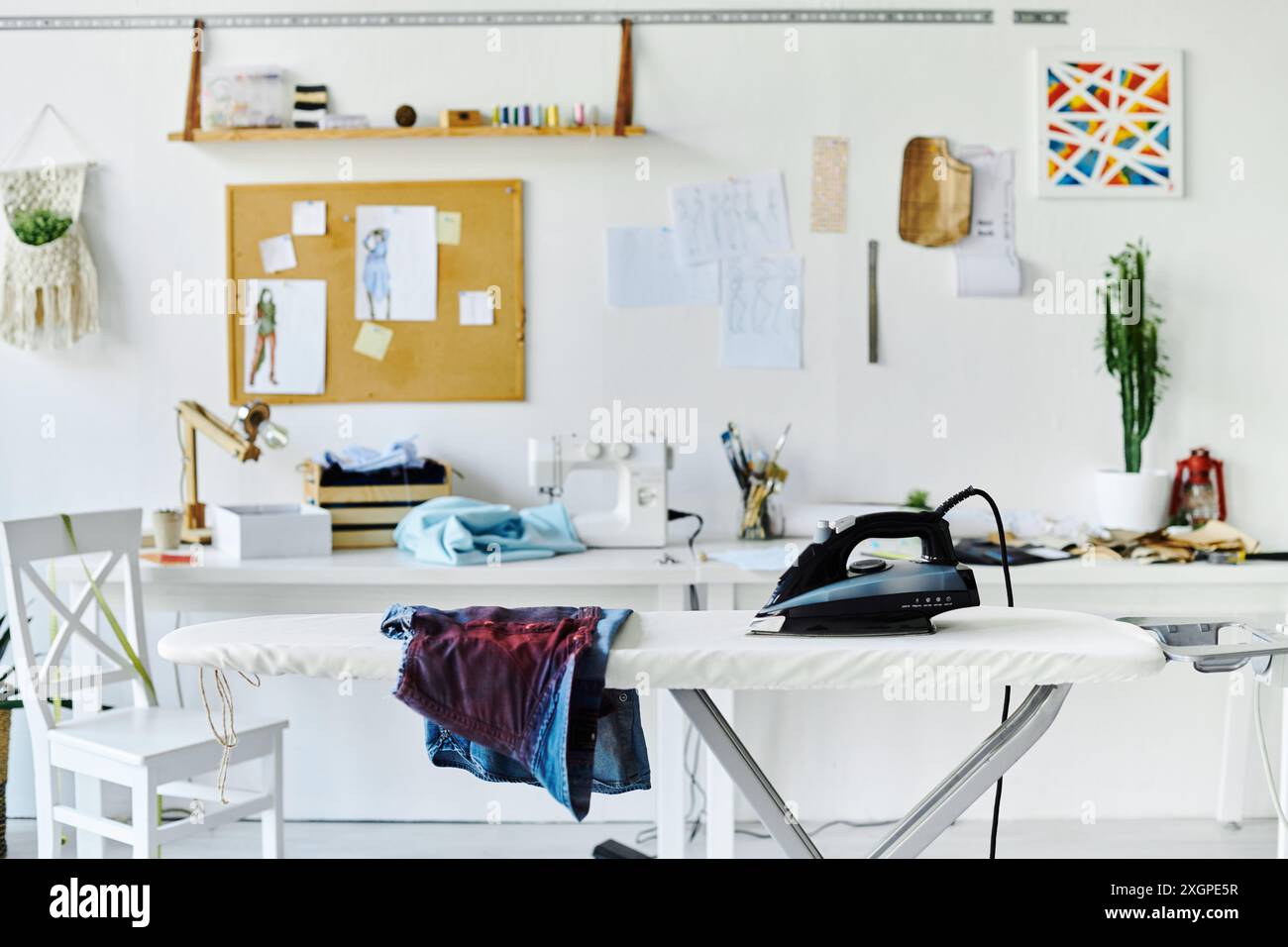 Un'area di lavoro pulita e ben illuminata, dotata di asse da stiro con indumenti disposti, ferro da stiro e macchina da cucire sullo sfondo. Foto Stock