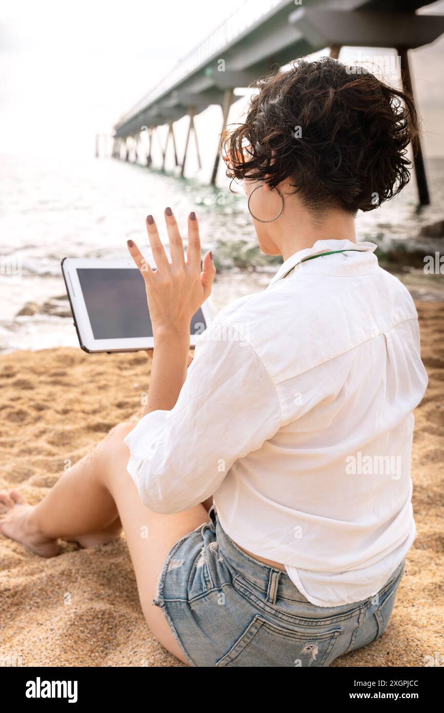 Ripresa verticale di una giovane donna che parla in videochiamata con atablet sulla spiaggia. Foto Stock