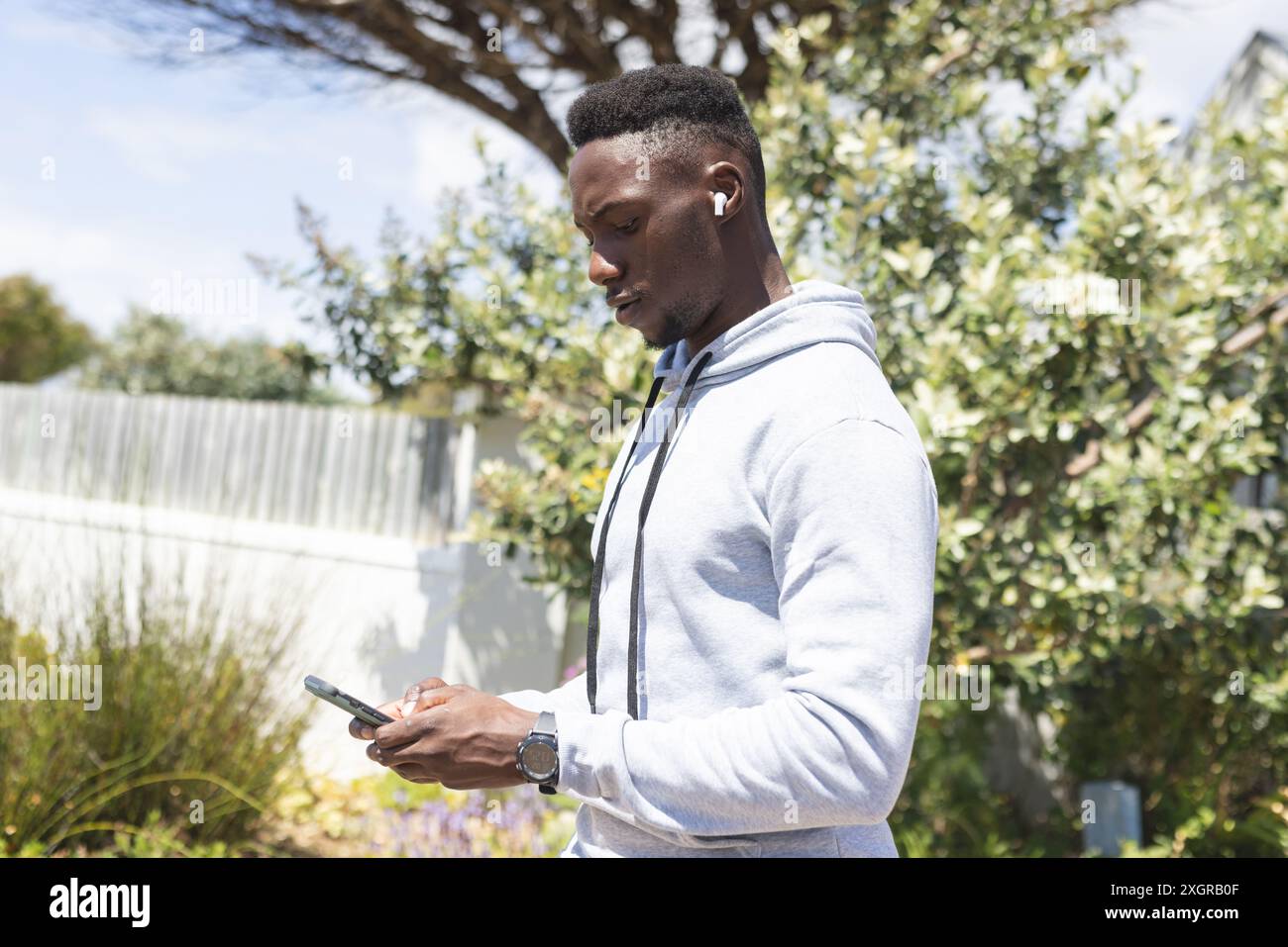 Uomo afroamericano concentrato che si sta allenando all'aperto, indossando auricolari e utilizzando smartphone. Comunicazione, jogging, fitness, esercizio fisico, benessere e guarigione Foto Stock