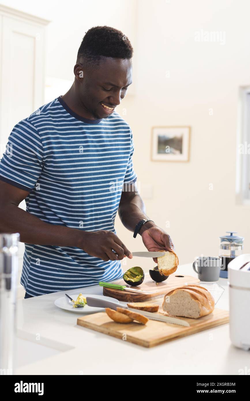 Uomo afroamericano sorridente che butta toast da avere con avocado in cucina. Cibo, cucina, vita domestica e stile di vita sano, inalterato. Foto Stock