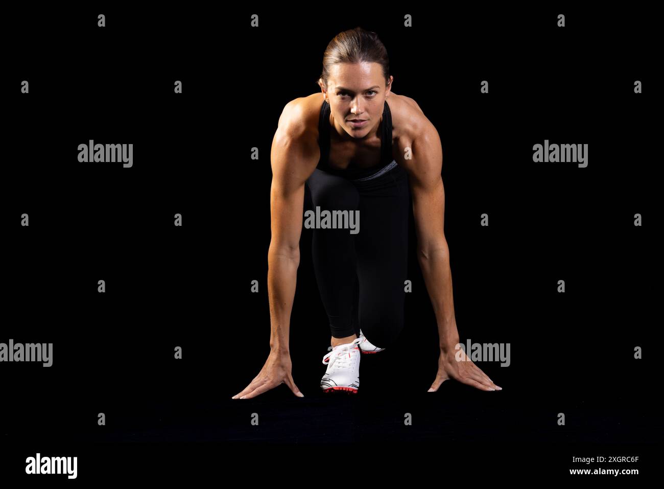 Giovane atleta caucasica nella posizione di partenza di un runner su sfondo nero, con spazio di copia. La sua intensa concentrazione esemplifica la determinazione e. Foto Stock