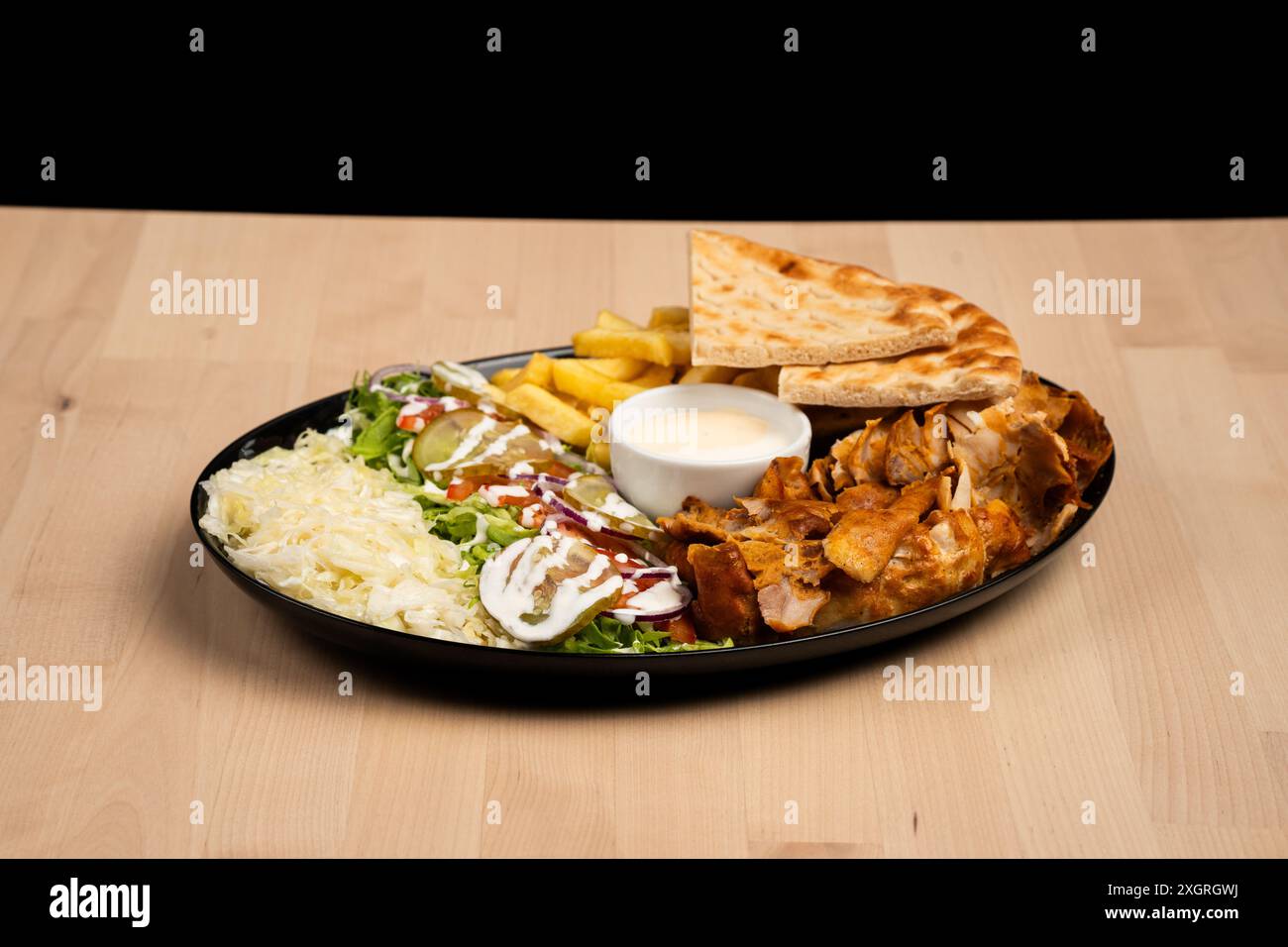 Un delizioso piatto di kebab doner misto con insalata, patatine fritte, pane pita e salsa su un tavolo di legno su sfondo nero Foto Stock