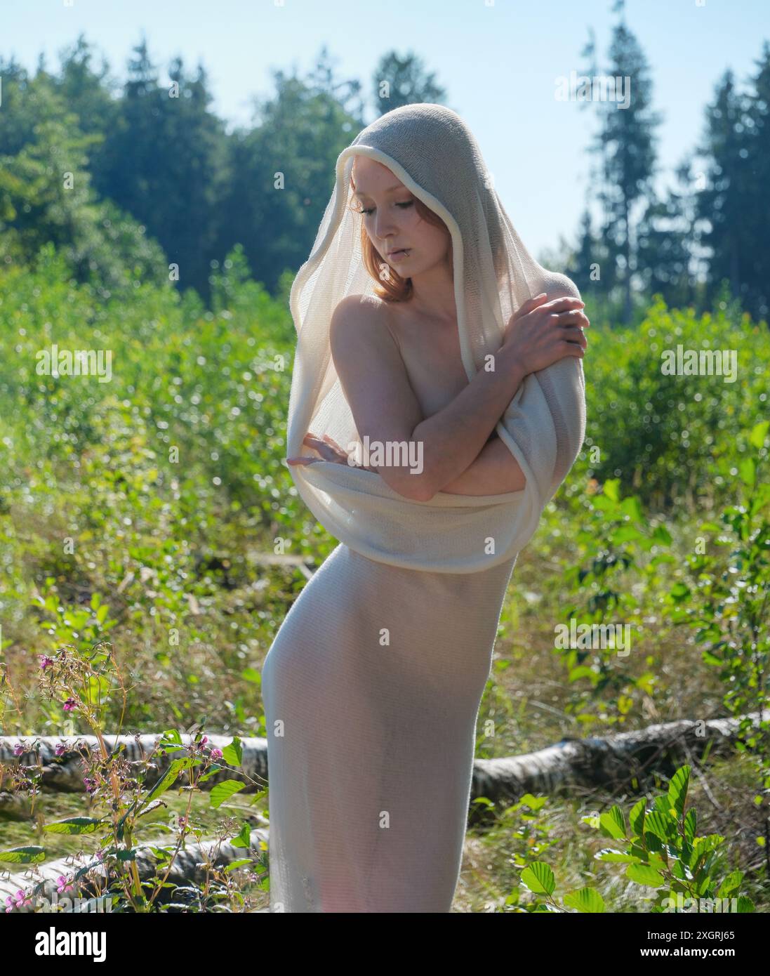 la donna sta aggraziatamente in mezzo alla natura, drappeggiata in un tessuto bianco puro. Guarda verso il basso, incarnando serenità ed eleganza, con vegetazione lussureggiante e sole Foto Stock