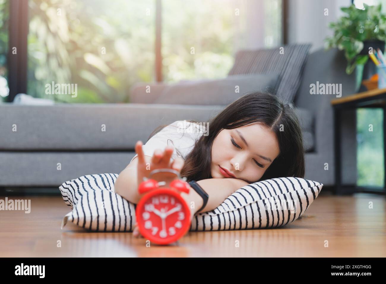 Giovane donna asiatica che si è rivolta a spegnere la sveglia mentre si trova sul pavimento nel soggiorno di casa. Foto Stock