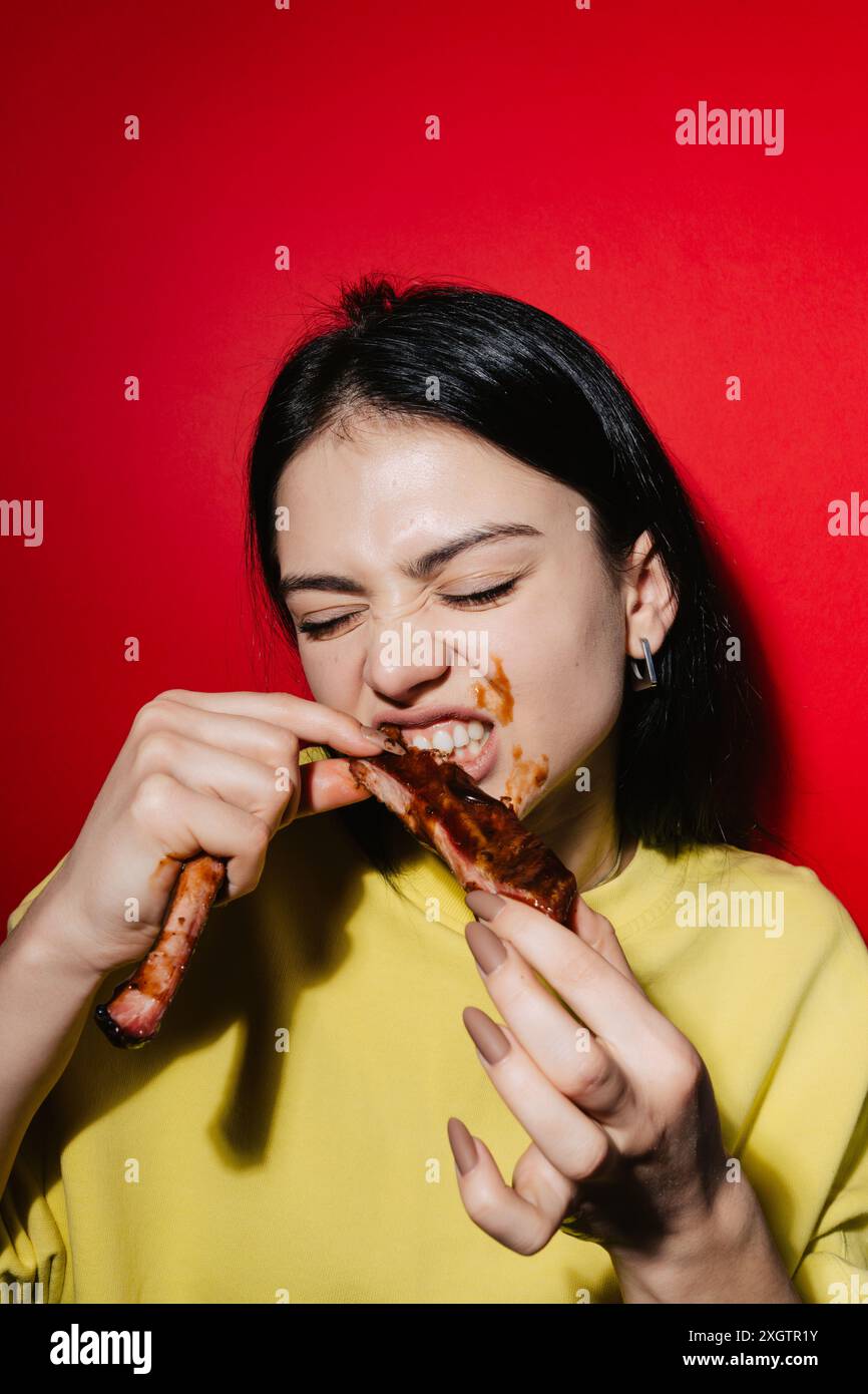 Un'immagine vibrante con una giovane donna in una camicia gialla, che mangia allegramente le costine del barbecue, le mani e le guance sono scherzosamente spalmate di salsa, set ag Foto Stock