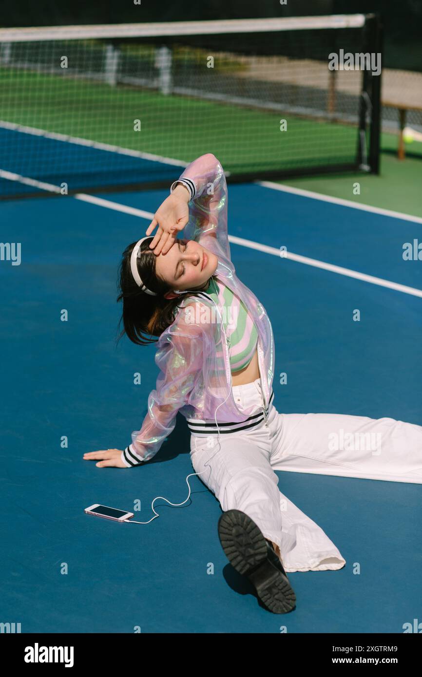 Foto vibrante di una giovane donna seduta su un campo da tennis, con gli occhi chiusi, indossa una giacca e ascolta la musica attraverso le cuffie collegate a uno smar Foto Stock