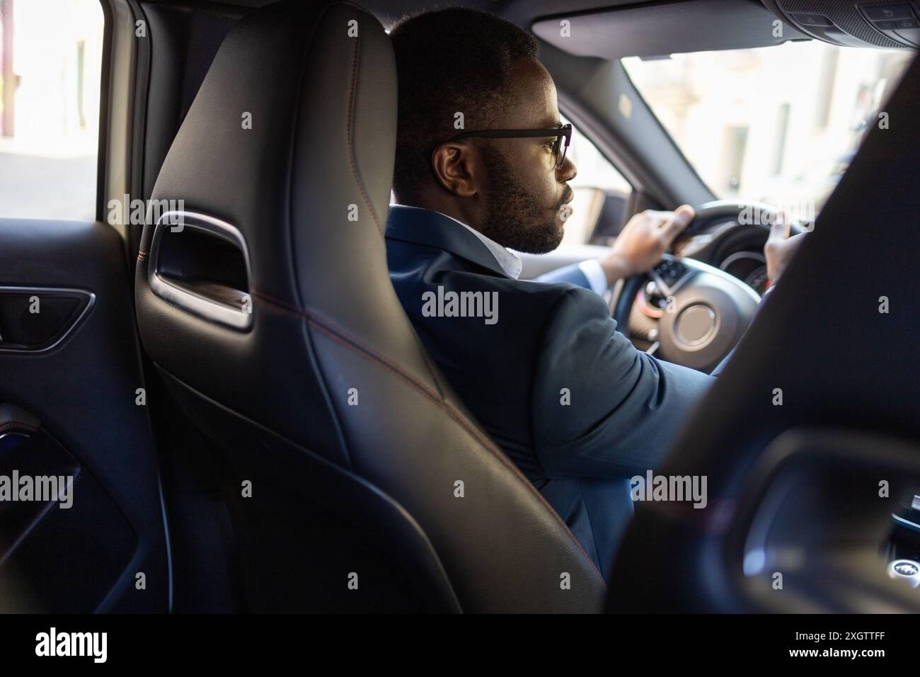 Uomo d'affari afroamericano che guarda lontano mentre guida un'auto di lusso. Indossa un vestito e degli occhiali. Foto Stock