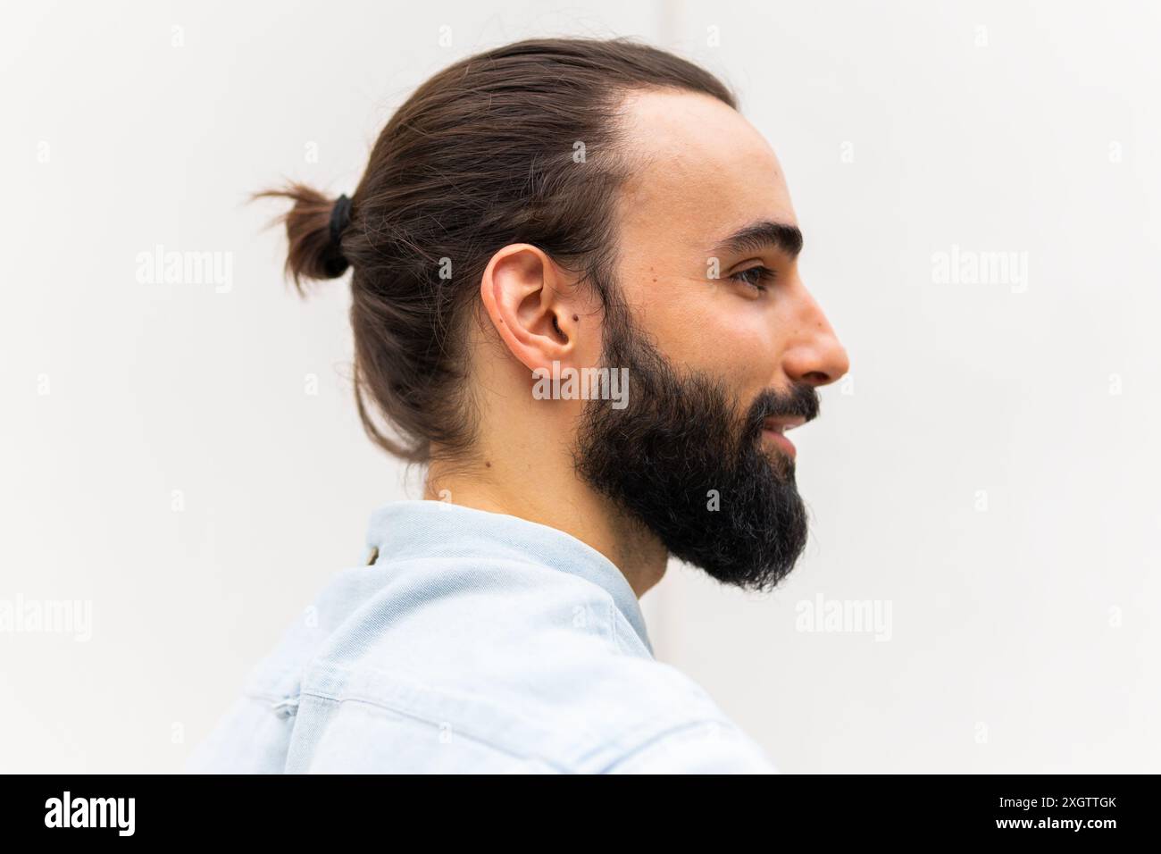 Profilo laterale di un giovane maschio adulto con una barba ordinata e una coda di cavallo, vestito con una camicia casual azzurra, che poggia su un bac semplice e di colore chiaro Foto Stock