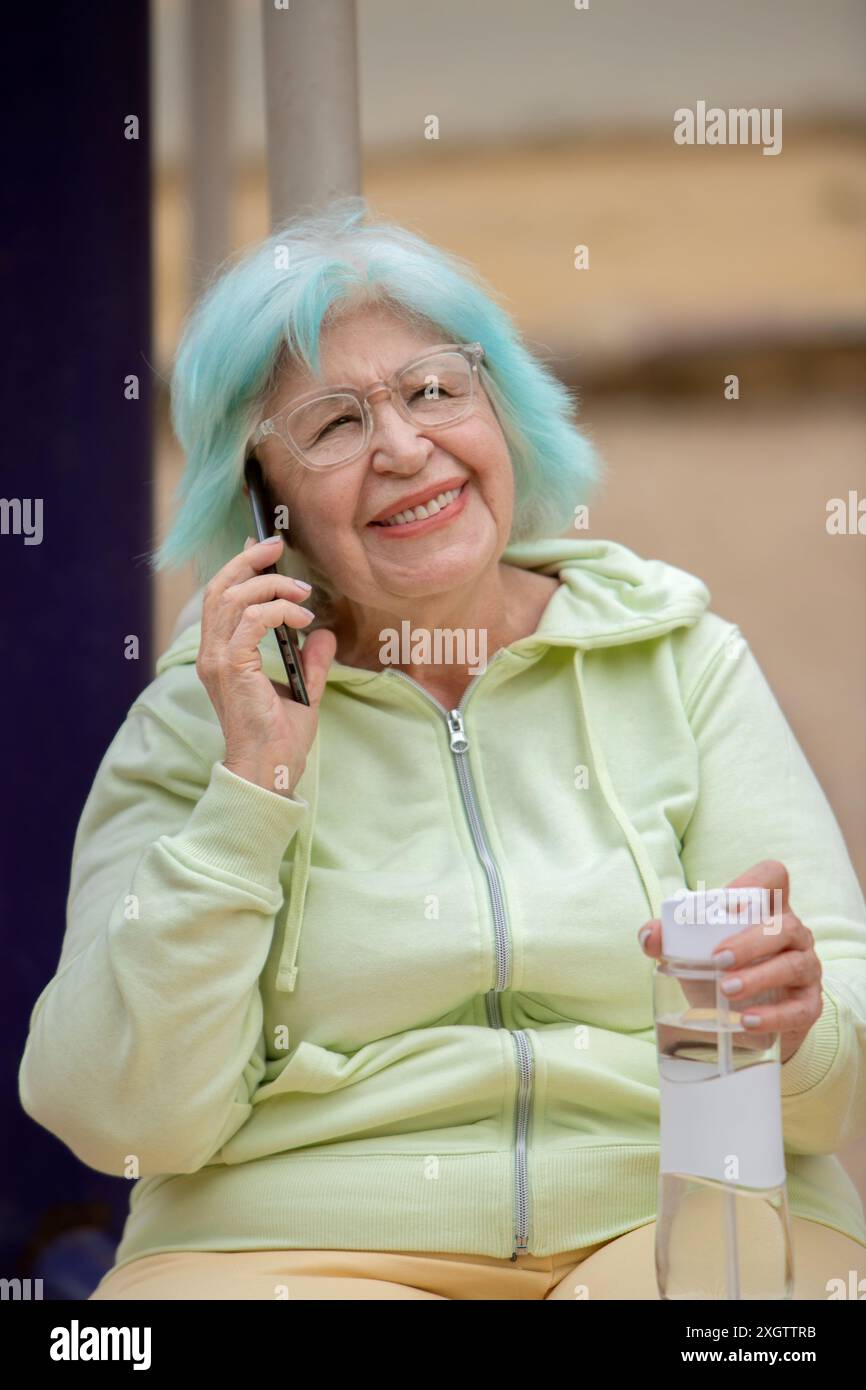 Donna anziana sorridente con capelli di tè unici utilizzando uno smartphone mentre è seduta nel parco. Ha in mano una bottiglia d'acqua e vestita con un abbigliamento sportivo, t Foto Stock