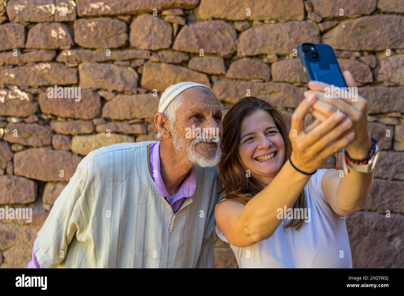 Un anziano marocchino e una giovane donna sorridono gioiosamente mentre scattano un selfie con uno smartphone contro un muro di pietra rustico in Marocco, Nort Foto Stock