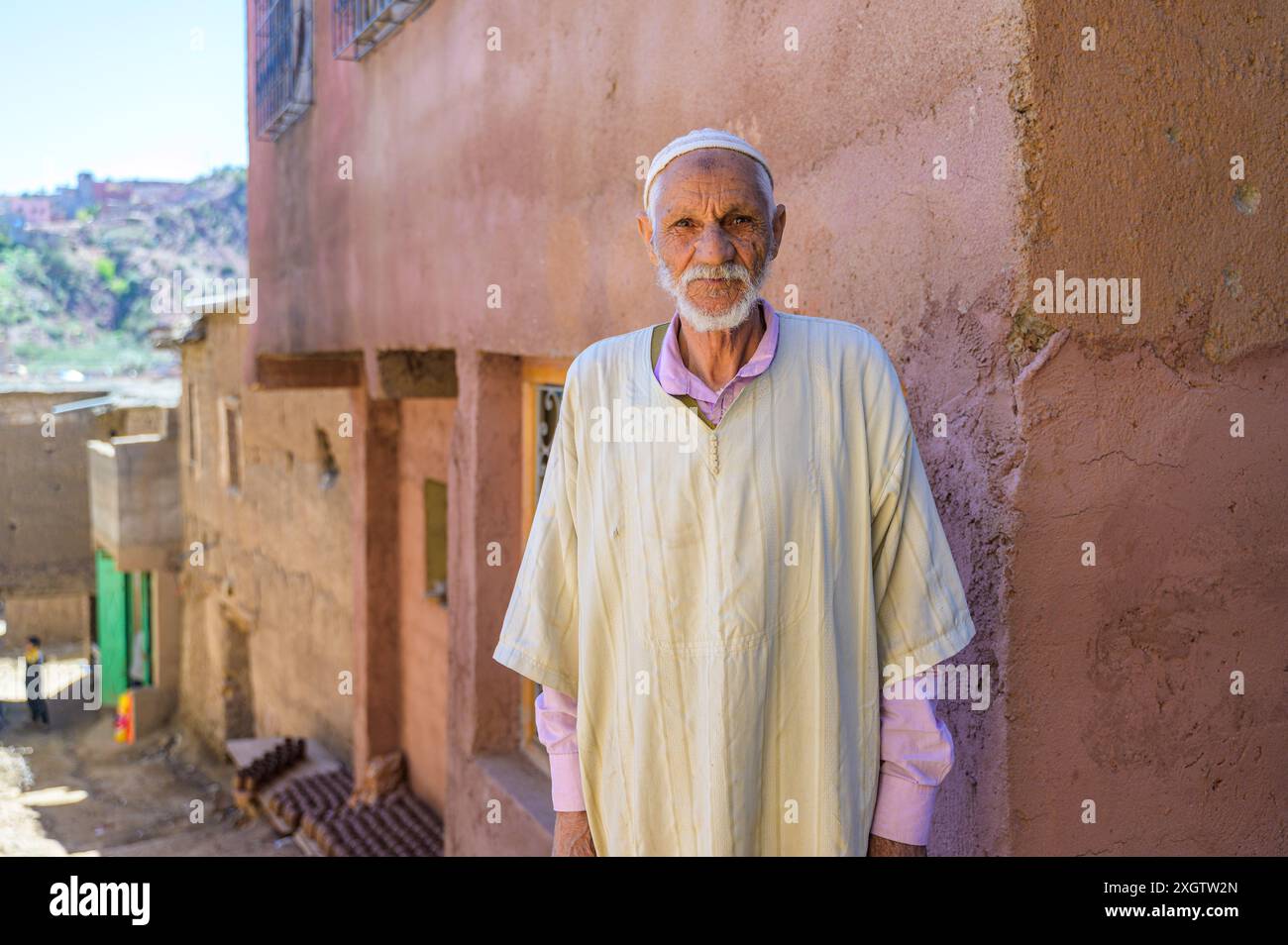 Un anziano uomo marocchino si erge con sicurezza davanti a una casa tradizionale del villaggio, indossando una classica djellaba bianca e una capsula, che incarna i ricchi c Foto Stock