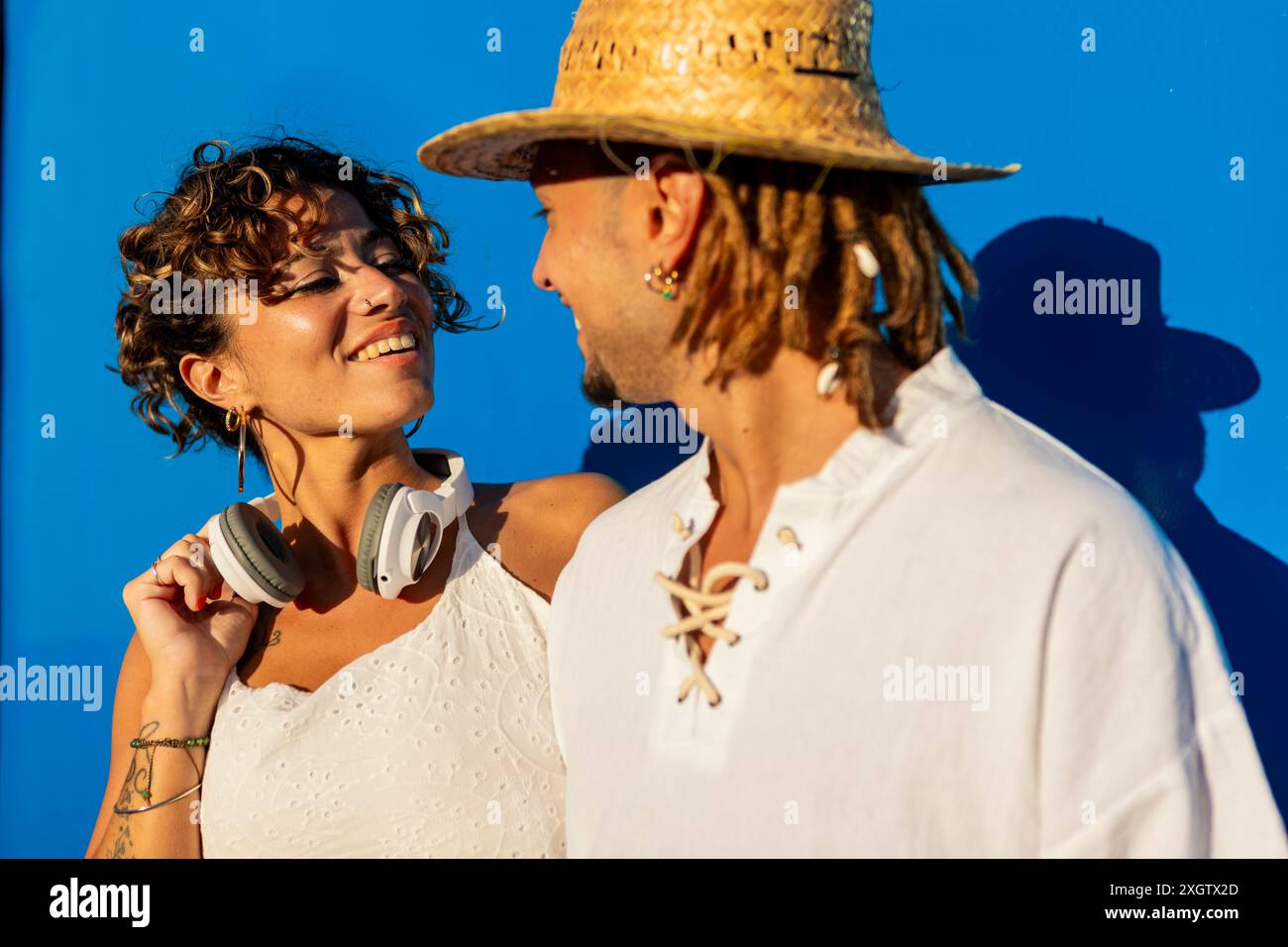 Un uomo e una donna caucasici gioiosi, che interagiscono su uno sfondo blu vivace. La donna con le cuffie guarda affettuosamente l'uomo con un cappello di paglia, Foto Stock