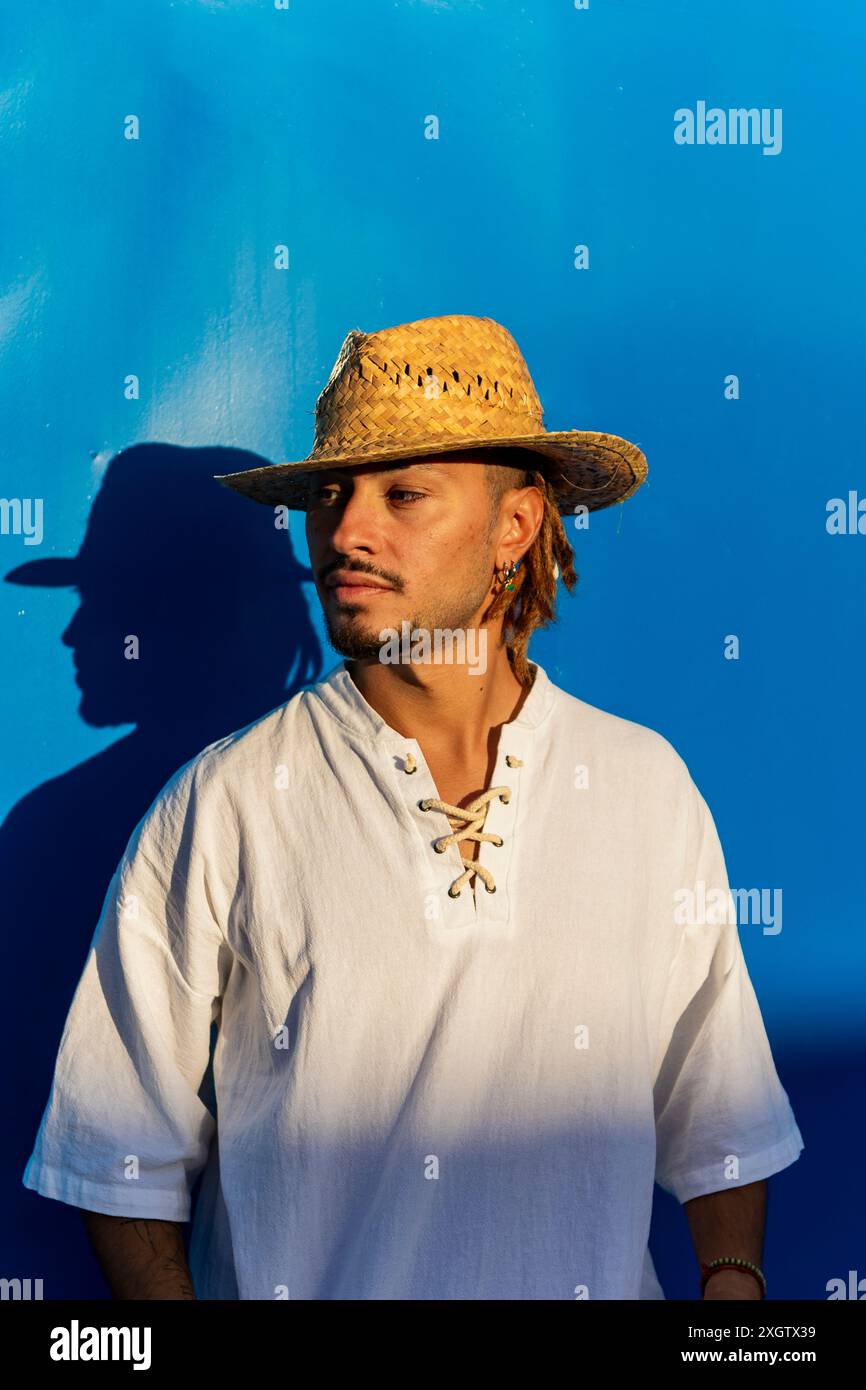 Un giovane uomo caucasico con dreadlock, con un cappello di paglia e una camicia stringata, guarda con attenzione su uno sfondo blu vivace. Foto Stock