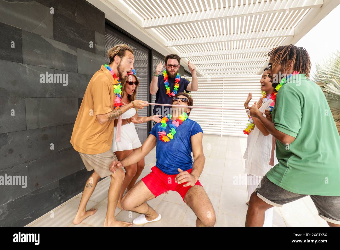 Gruppo multietnico di amici che giocano con entusiasmo a un gioco giocoso durante una riunione estiva, indossando colorate leis floreali ed esprimendo gioia. Foto Stock