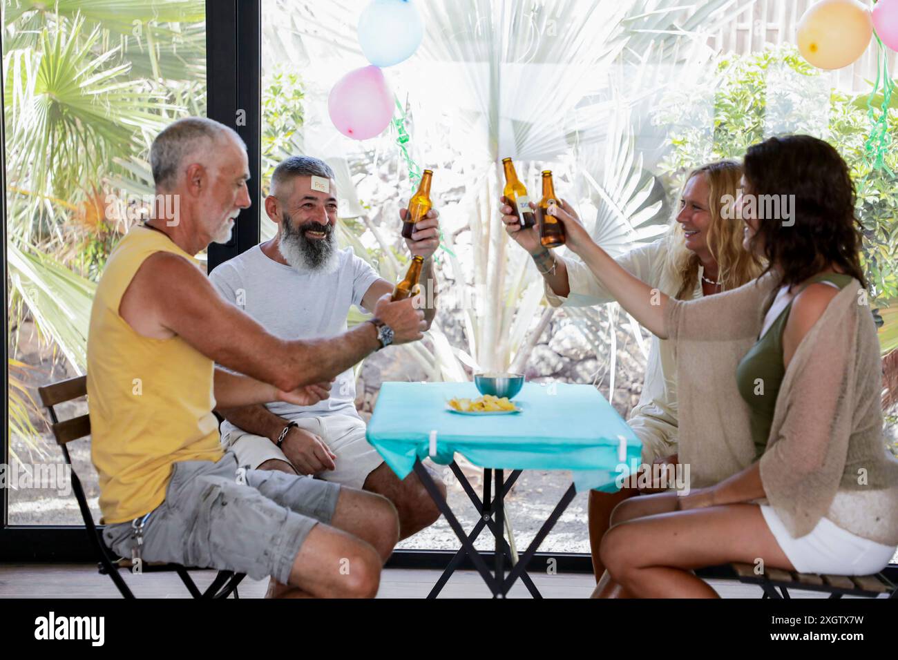 Un gruppo di amici anziani si riunisce all'aperto, brindando con delle birre sotto un riparo decorato con palloncini, circondato da una vegetazione lussureggiante e godendosi la luce Foto Stock