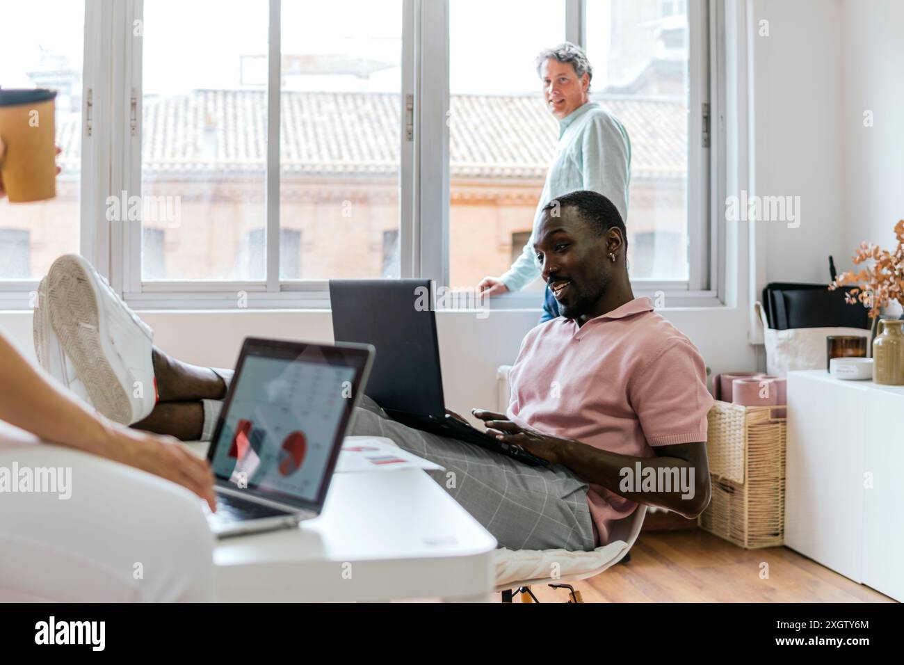 In un ambiente di coworking illuminato dal sole, un afroamericano che digita su un laptop guarda lo schermo mentre un anziano uomo caucasico si trova nel backgroun Foto Stock