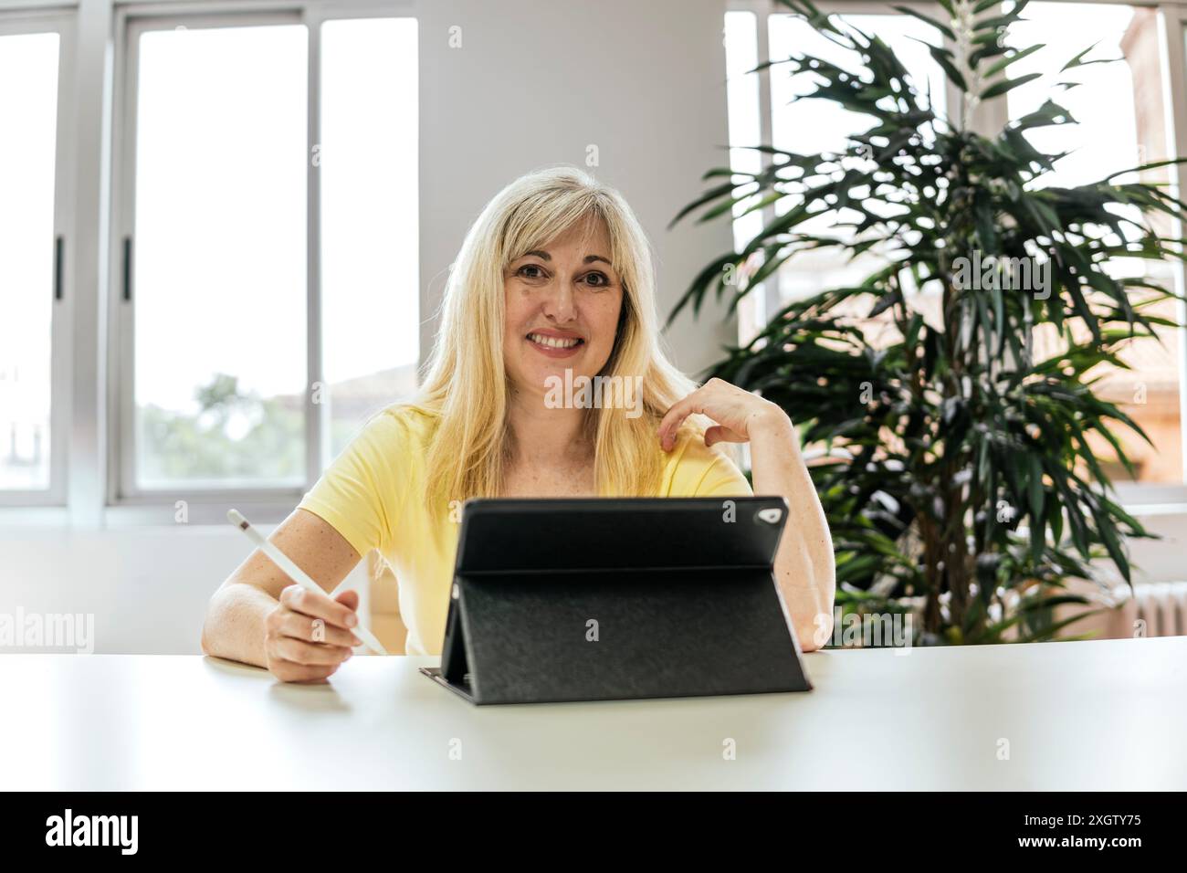 Una donna caucasica sorride alla fotocamera mentre lavora su un tablet in uno spazio di coworking ben illuminato, circondato da vivaci piante interne. Foto Stock