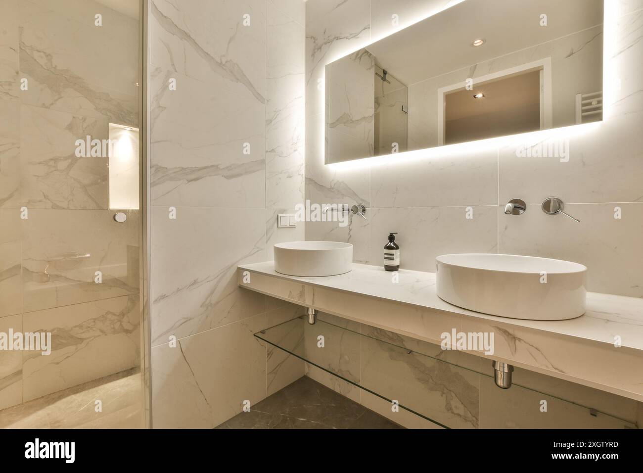 Un bagno contemporaneo presenta eleganti pareti e ripiani in marmo bianco con due eleganti lavandini rotondi, impianti moderni e illuminazione delicata. Foto Stock