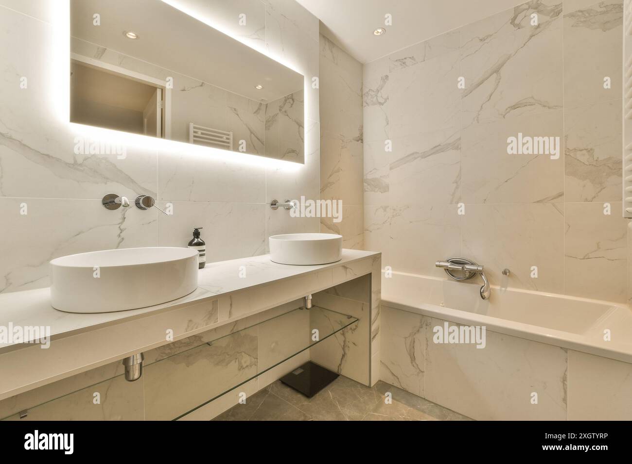 Il bagno elegante e moderno presenta pareti e ripiani in marmo, due lavandini, uno specchio retroilluminato e finiture minimaliste. Foto Stock