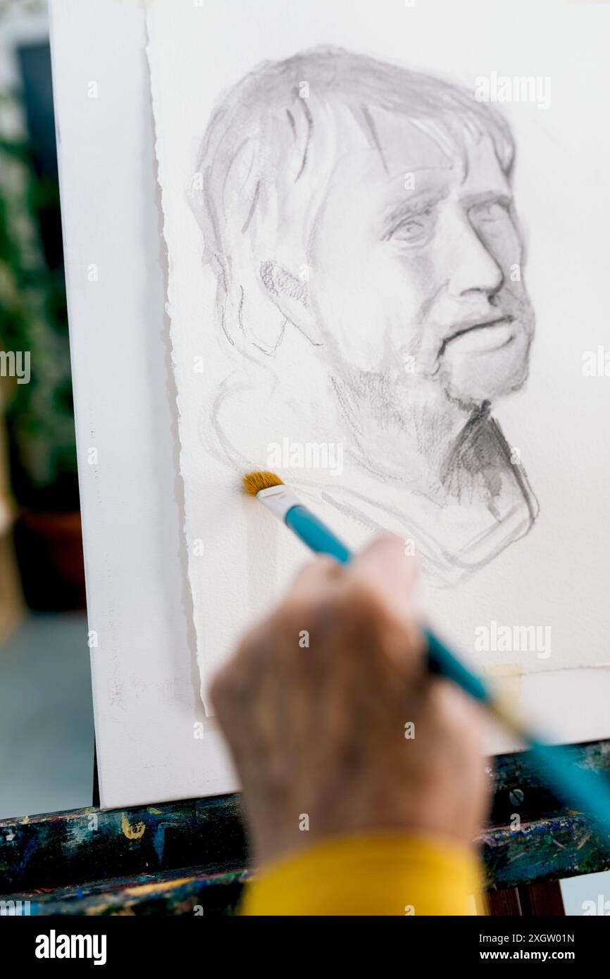 Una donna anziana anonima dipinge meticolosamente un ritratto maschile dettagliato, concentrandosi sul rendering delle caratteristiche del viso con abilità e precisione. La sua mano che tiene Foto Stock