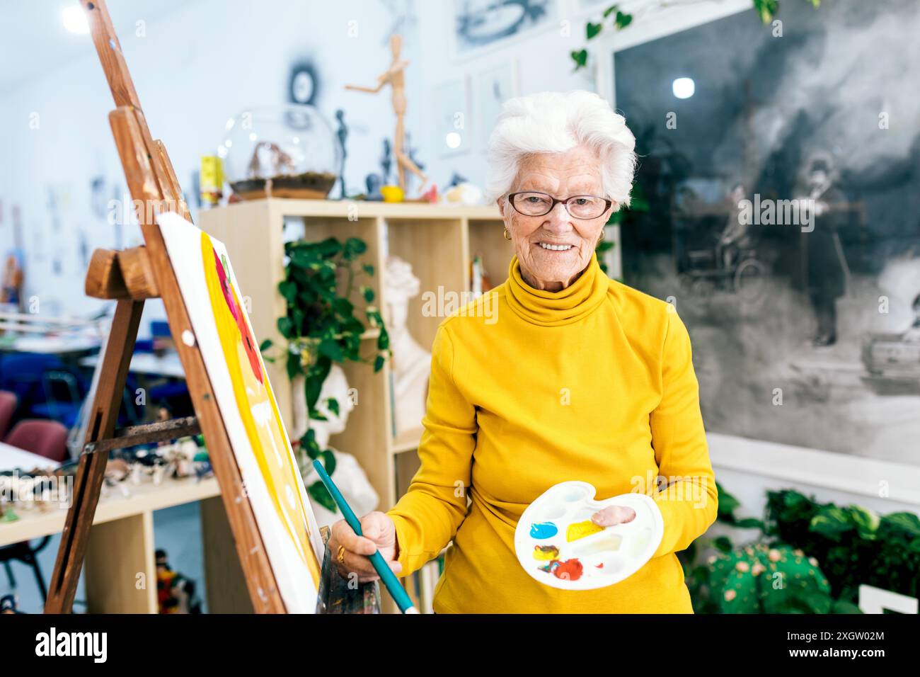 Una donna anziana allegra con i capelli bianchi dipinge su una tela in uno studio d'arte illuminato, tenendo in mano una tavolozza e una spazzola Foto Stock