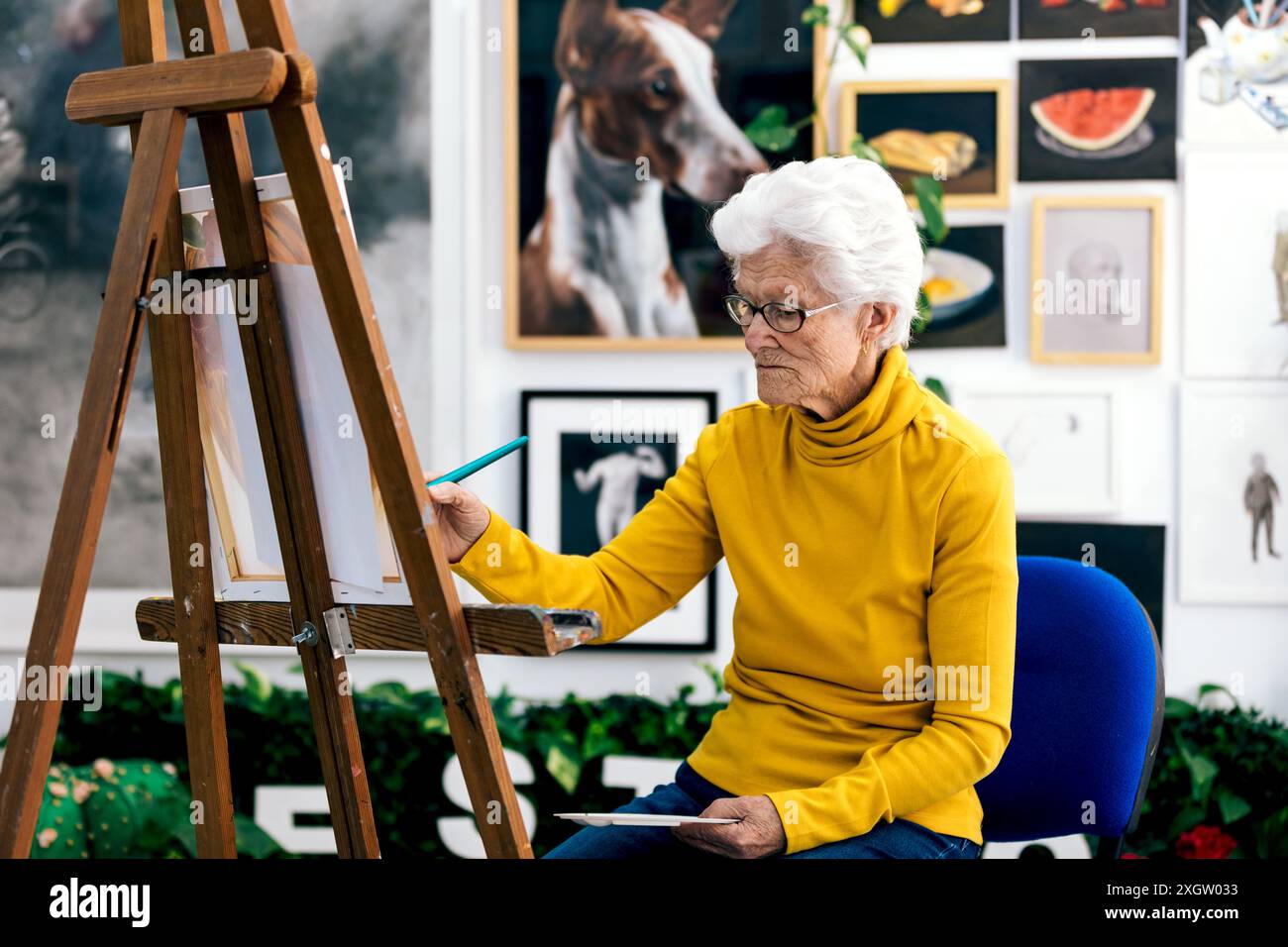 Una donna anziana, situata in un vivace studio d'arte pieno di varie opere d'arte, dipinge con attenzione su una tela. Indossa un top giallo brillante, complementi Foto Stock