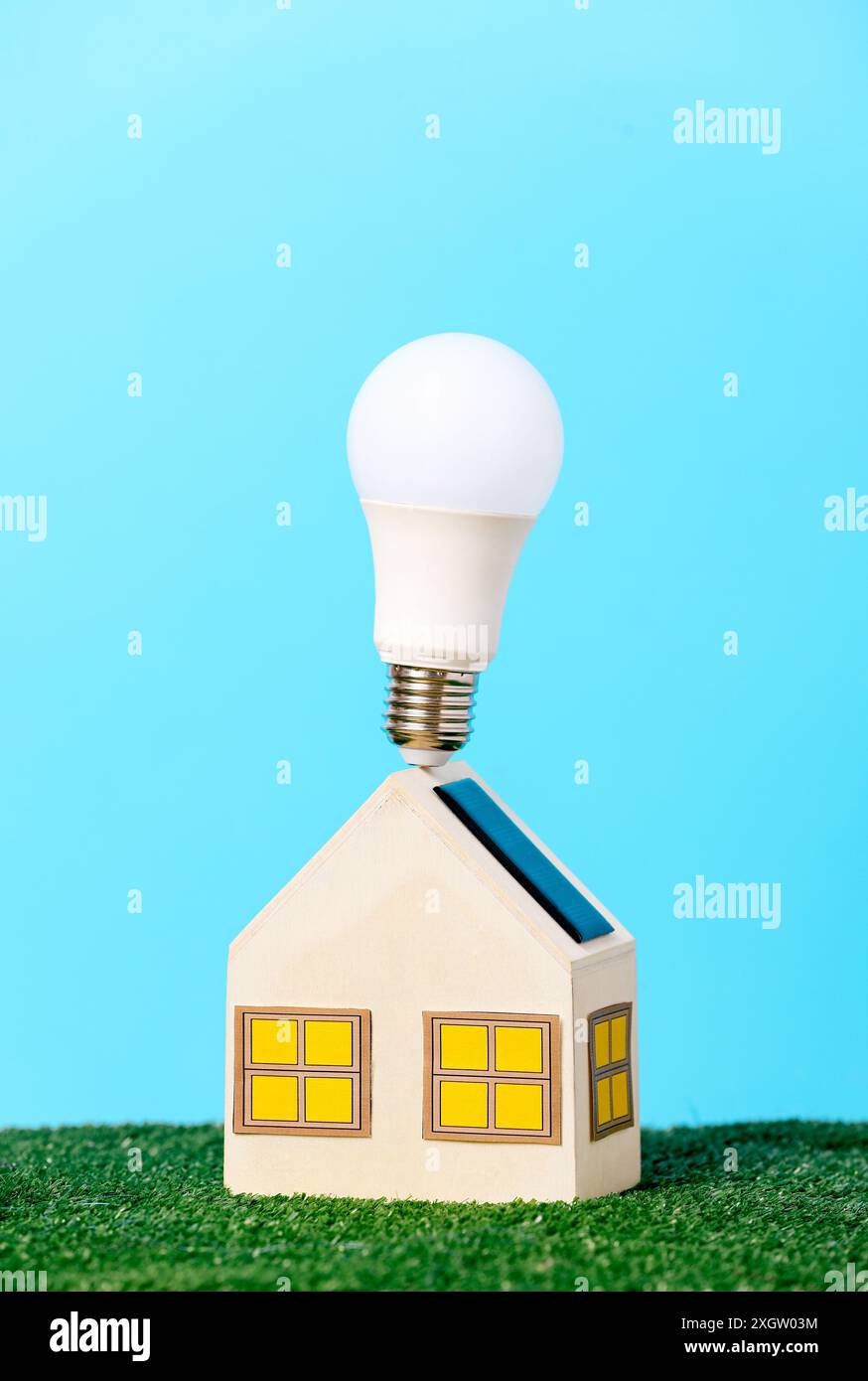 Un mini modello di casa in legno coronato da una lampadina a LED ecocompatibile che simboleggia soluzioni di alloggiamento sostenibili, posizionato su una lussureggiante erba verde con una vivace b Foto Stock