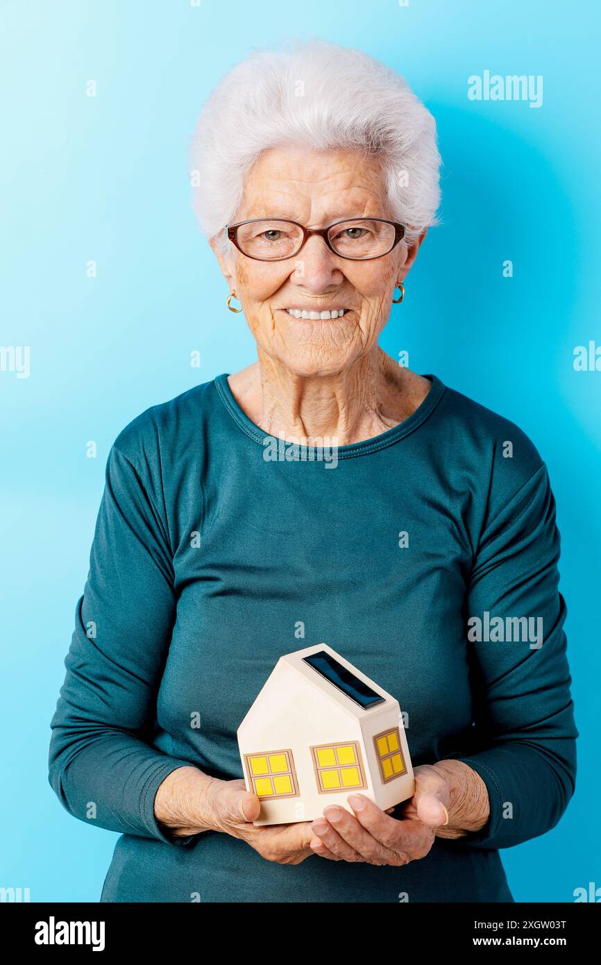 Una donna anziana allegra con i capelli bianchi, con occhiali e una camicia color verde acqua scura, è orgogliosa di ospitare un piccolo modello di casa dotato di pannello solare, S Foto Stock
