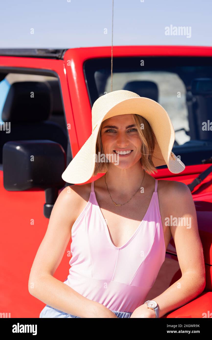 La giovane donna caucasica posa di fronte a un veicolo rosso durante un viaggio in auto. Il suo vestito estivo e il suo sorriso luminoso suggeriscono una piacevole avventura all'aria aperta. Foto Stock