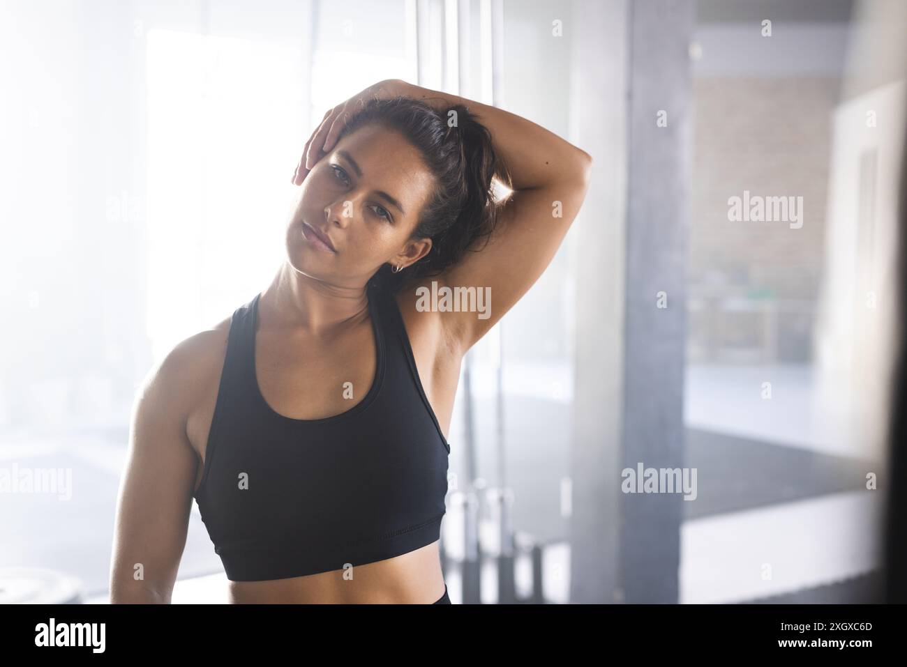 Giovane donna birazziale posa con fiducia in palestra, trasudando forza e fitness. Il suo abbigliamento da allenamento e la sua posizione suggeriscono una pausa nella sua forma fisica rigorosa Foto Stock