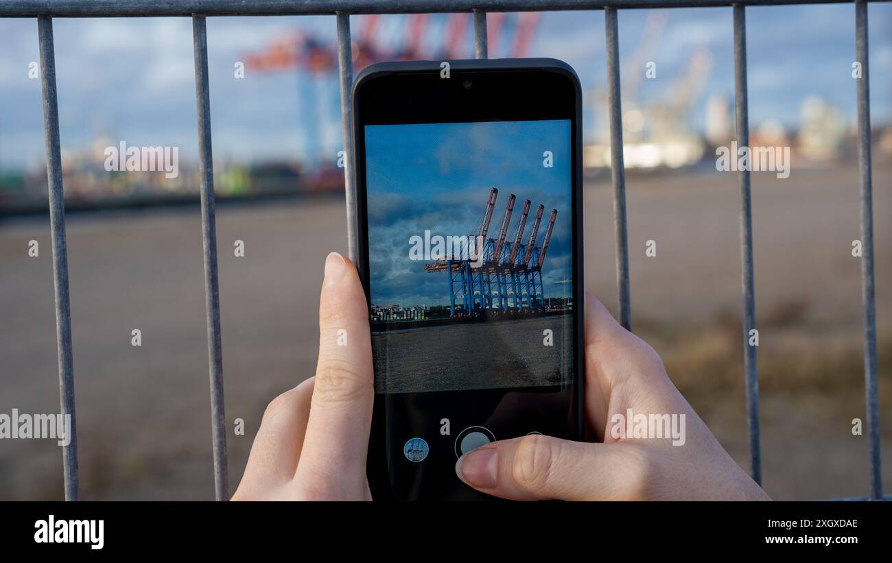 Una donna tiene in mano il suo smartphone e scatta una foto di un porto di spedizione mentre si trova dietro una recinzione Foto Stock