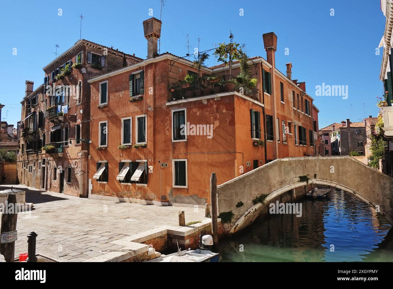 Palazzo Veneziano rosso con patio, piante in vaso, classico camino veneziano a cono invertito, campo con pozzo comune e ponte in muratura sul canale Foto Stock