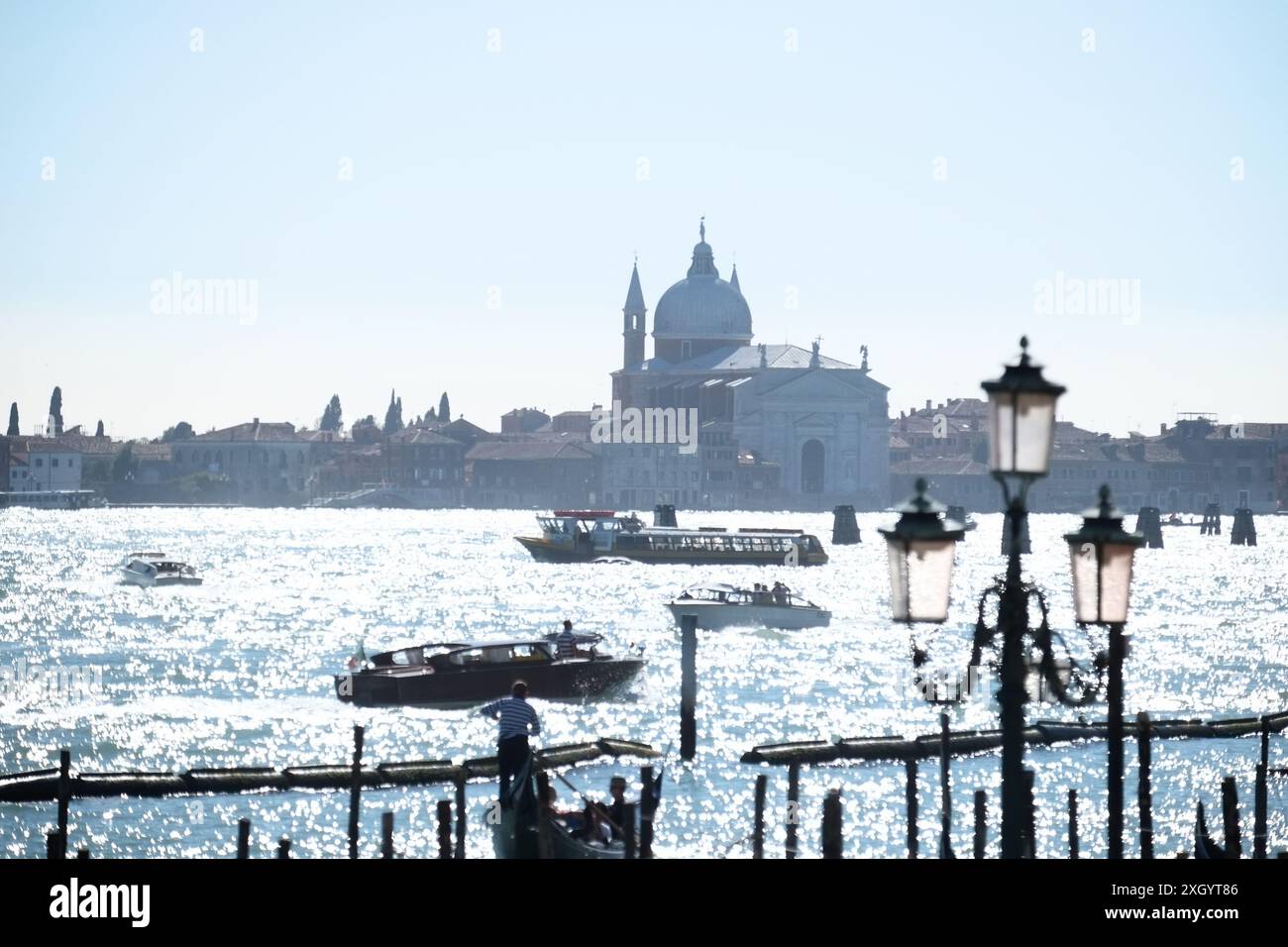 Barche che attraversano la scintillante distesa d'acqua tra Riva degli Schiavoni e la Chiesa del Santissimo Redentore incorniciata da una lampada e da un cielo azzurro soffice Foto Stock