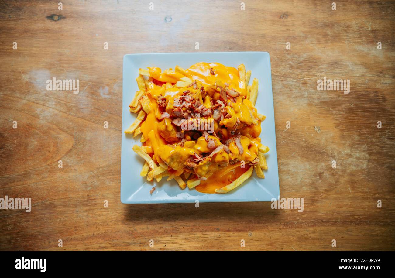 Vista dall'alto delle patatine fritte ricoperte di formaggio cheddar e pancetta su un tavolo di legno. Patatine fritte immerse in formaggio giallo con pancetta su tavola di legno Foto Stock