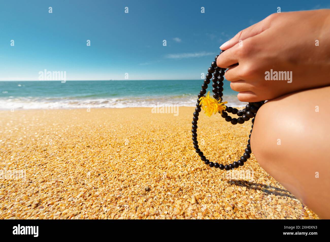 La mano di una donna bianca regge un rosario per la pratica della preghiera e della meditazione sullo sfondo della costa sabbiosa del mare e del cielo. All'aperto Foto Stock