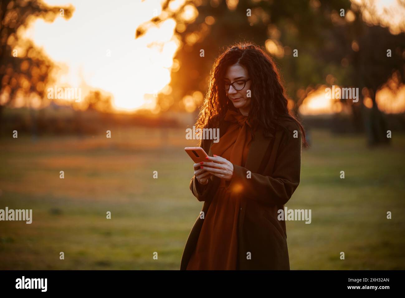 Una donna con lunghi capelli ricci che indossa un cappotto marrone e un abito color ruggine usa il suo smartphone mentre si trova in un parco con un sole tramontare alle spalle. Foto Stock