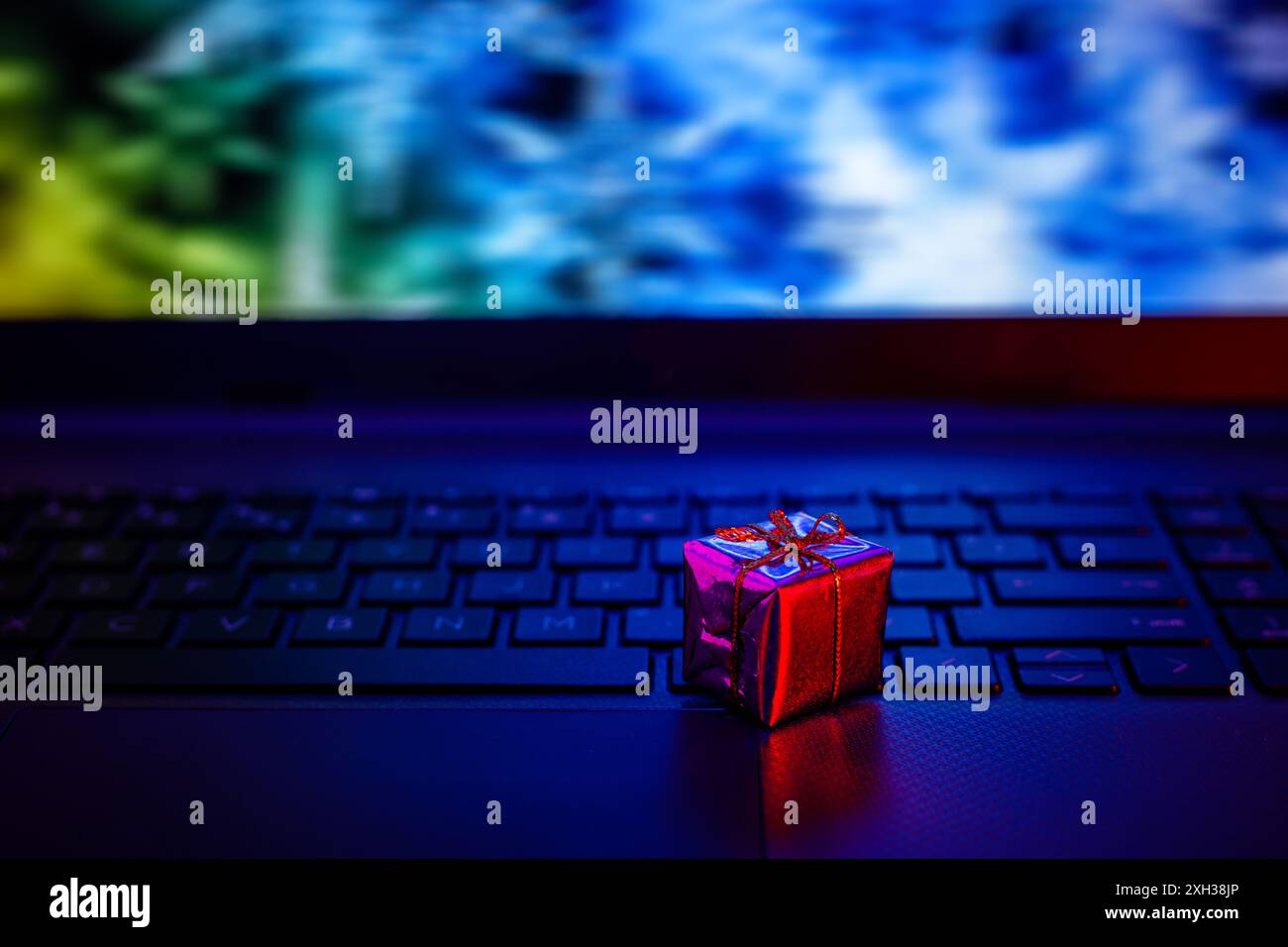 Una piccola confezione regalo rossa con fiocco dorato si trova su una tastiera nera per laptop. Lo schermo del notebook è fuori fuoco e mostra colori blu e verde. Foto Stock