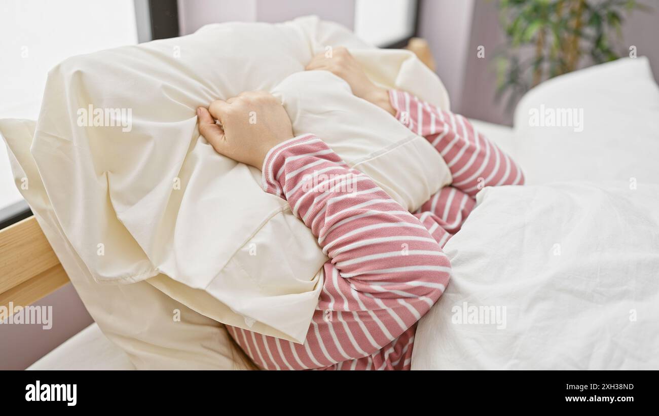 Una giovane donna in un pigiama a righe abbraccia un cuscino in un'accogliente camera da letto, evocando una sensazione di comfort e relax. Foto Stock