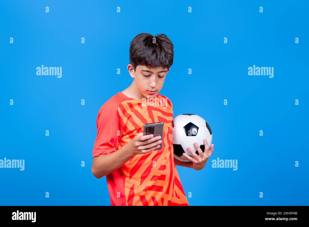 Un ragazzo con una maglietta arancione tiene in mano una palla da calcio e guarda il suo telefono. Concetto di sport e tecnologia. Foto Stock