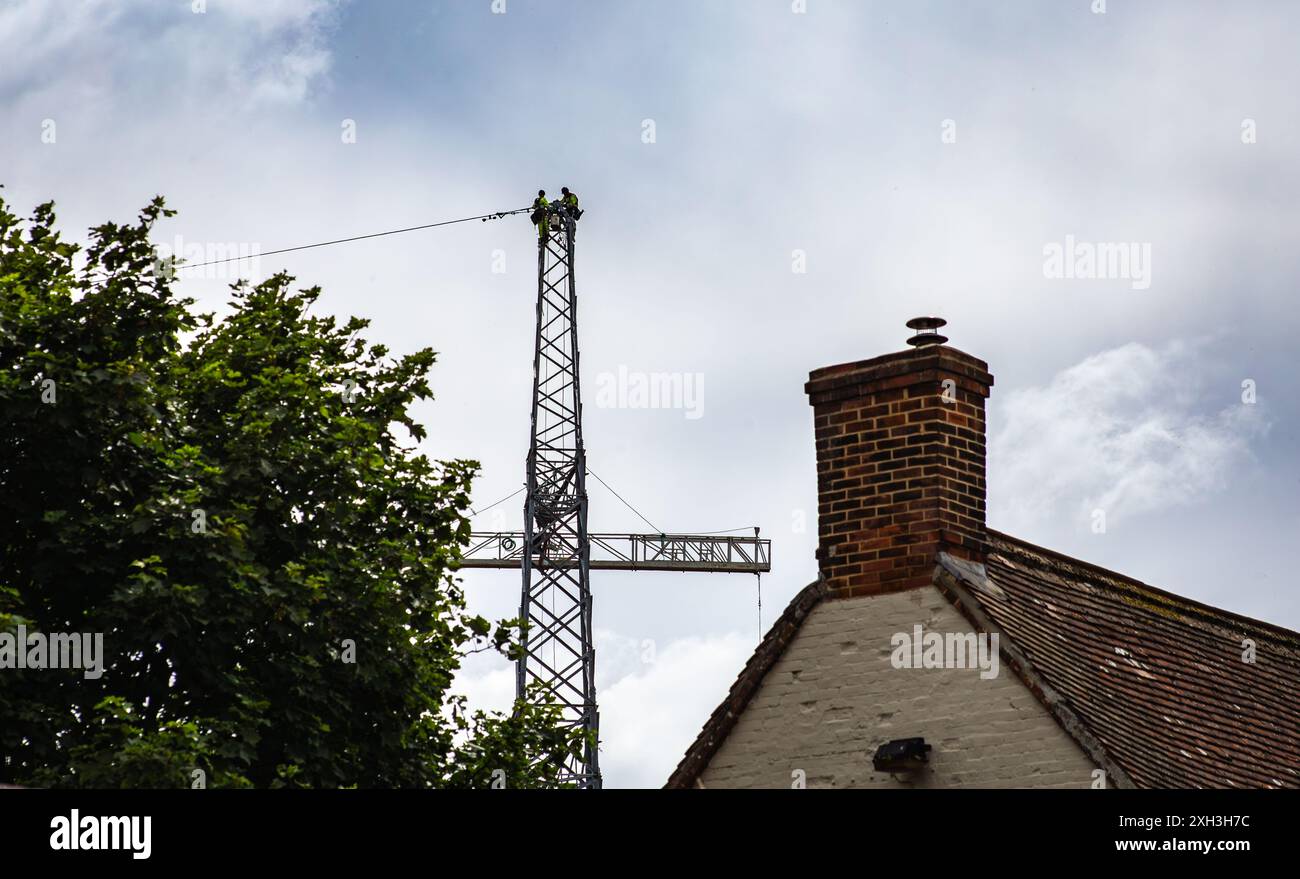 Edificio con un classico camino in mattoni adagiato contro un cielo nuvoloso, con due persone che lavorano in cima a un pilone elettrico in lontananza. Foto Stock