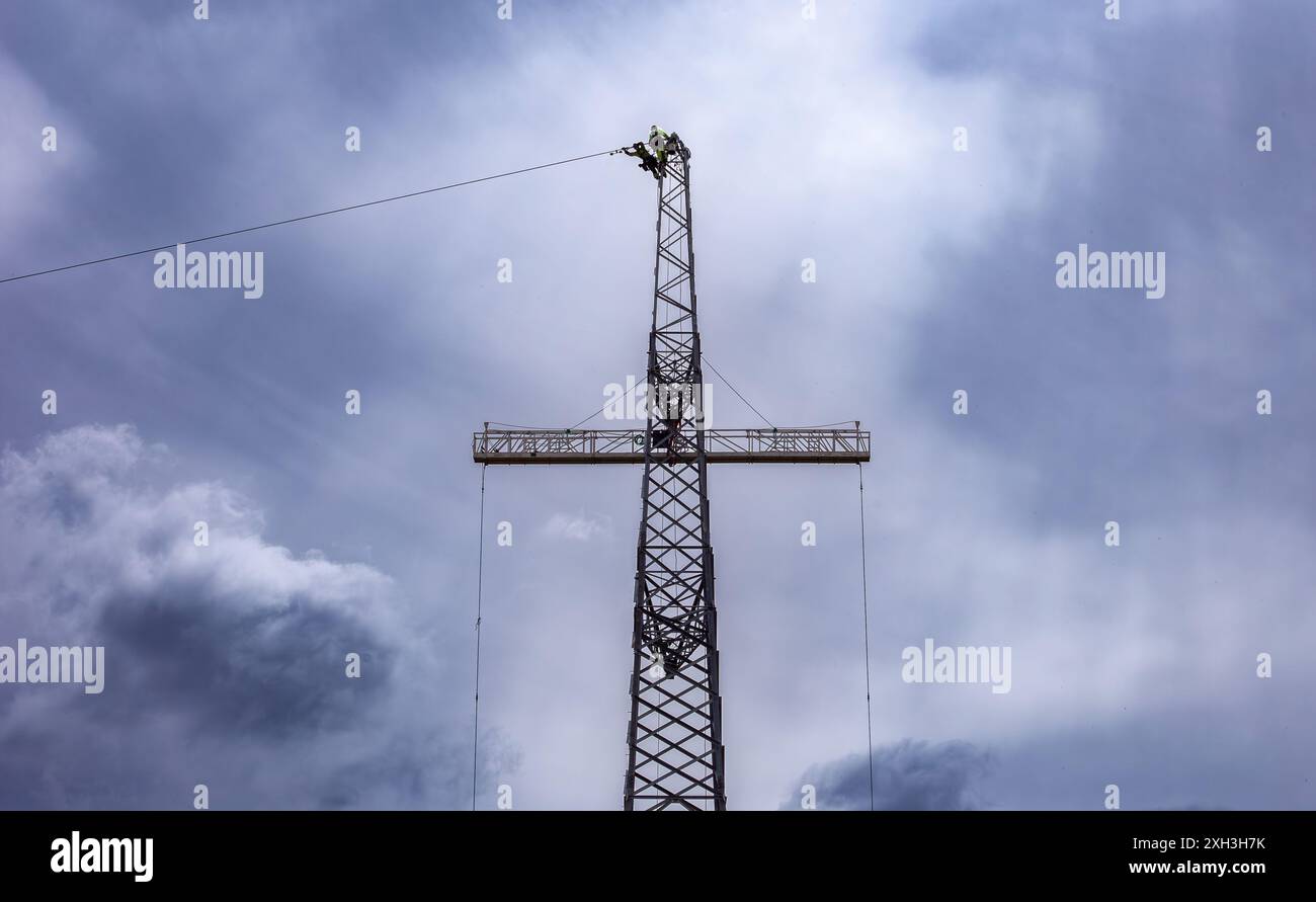 Lavoratore in indumenti ad alta visibilità in cima a una torre di pilastri elettrici contro un cielo nuvoloso con l'attrezzatura che viene avvolta su un cavo Foto Stock