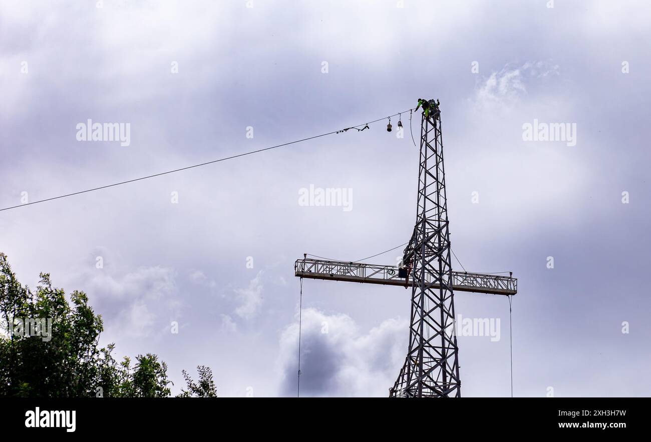 Lavoratore in indumenti ad alta visibilità in cima a una torre di pilastri elettrici contro un cielo nuvoloso con l'attrezzatura che viene avvolta su un cavo Foto Stock