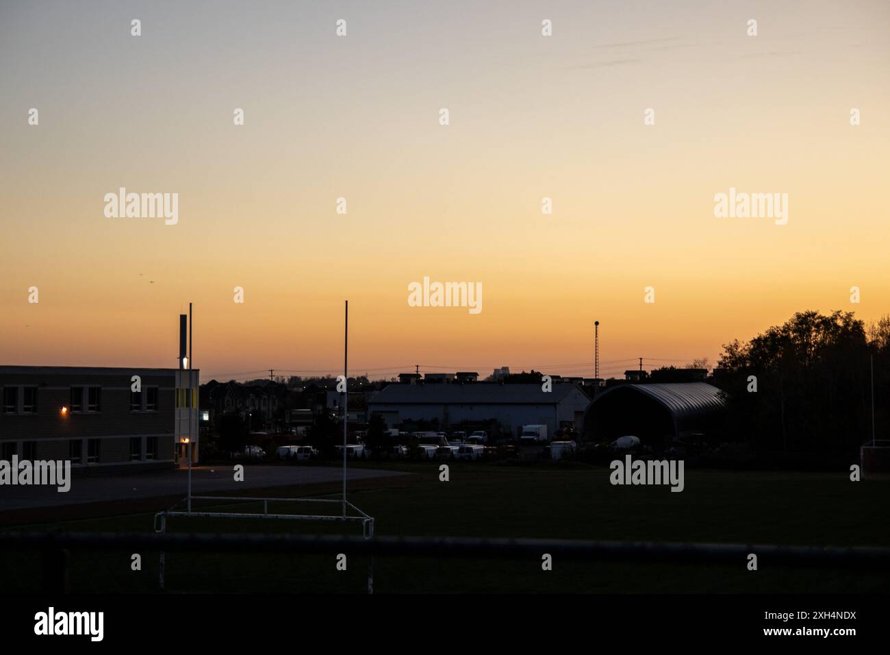 Caldo cielo arancione al crepuscolo - silhouette di pali di porta su un campo sportivo - edifici industriali delineano l'orizzonte - atmosfera serena. Preso in T Foto Stock