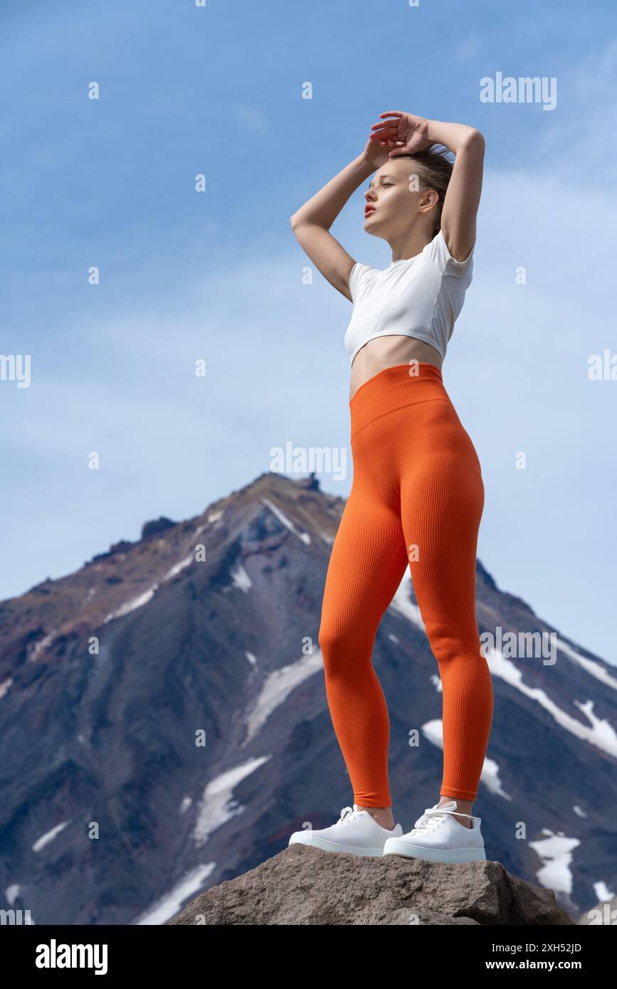 Istruttore di fitness slim con T-shirt, pantaloni da yoga arancioni e allenatori in piedi sullo sfondo del vulcano. Donna adulta in relax posa con le braccia Foto Stock